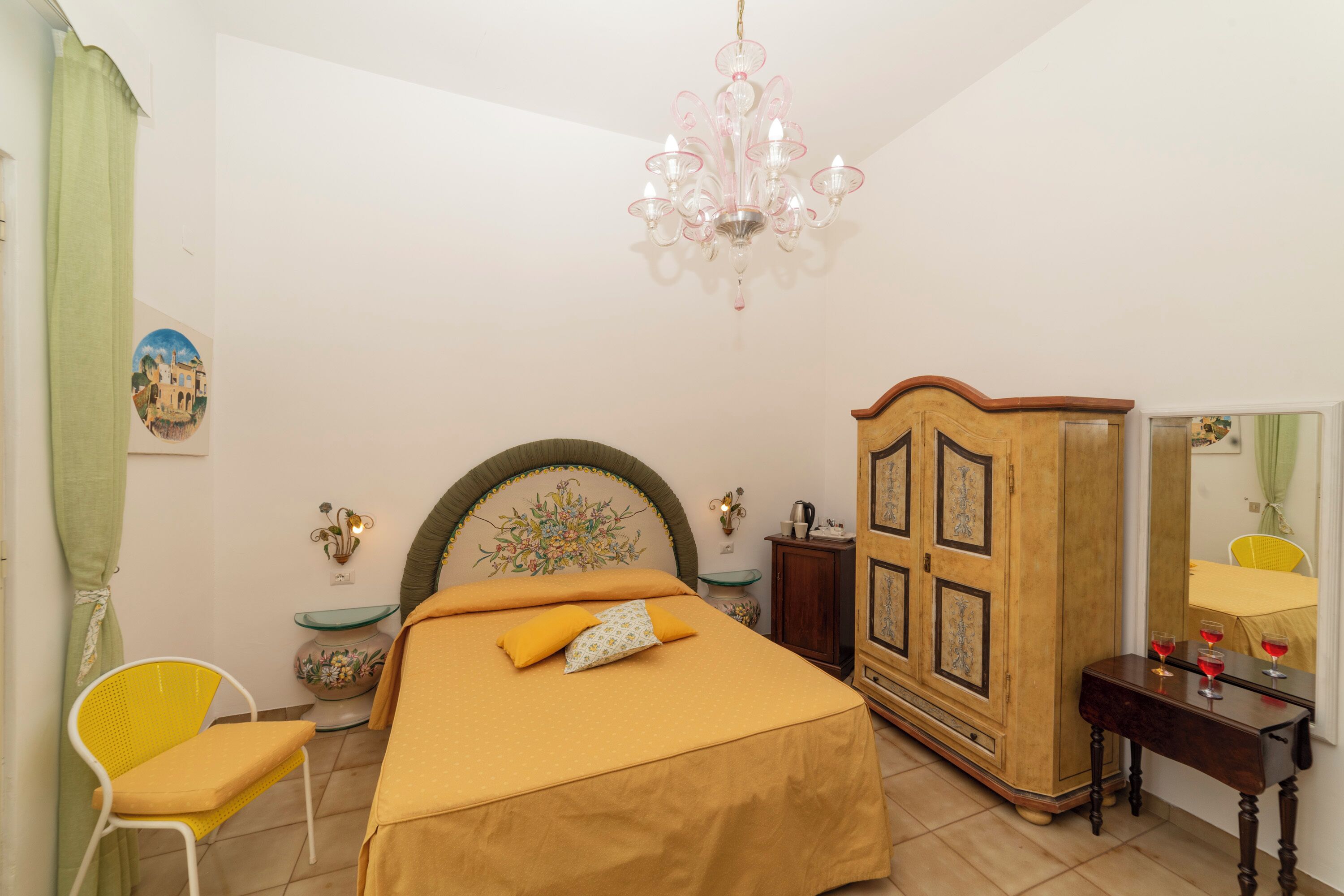 A bedroom with a bed, a wooden wardrobe, a chair, a table, a chandelier, and a mirror.