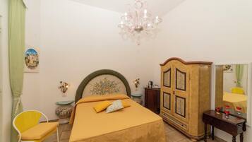 A bedroom with a bed, a wooden wardrobe, a chair, a table, a chandelier, and a mirror.