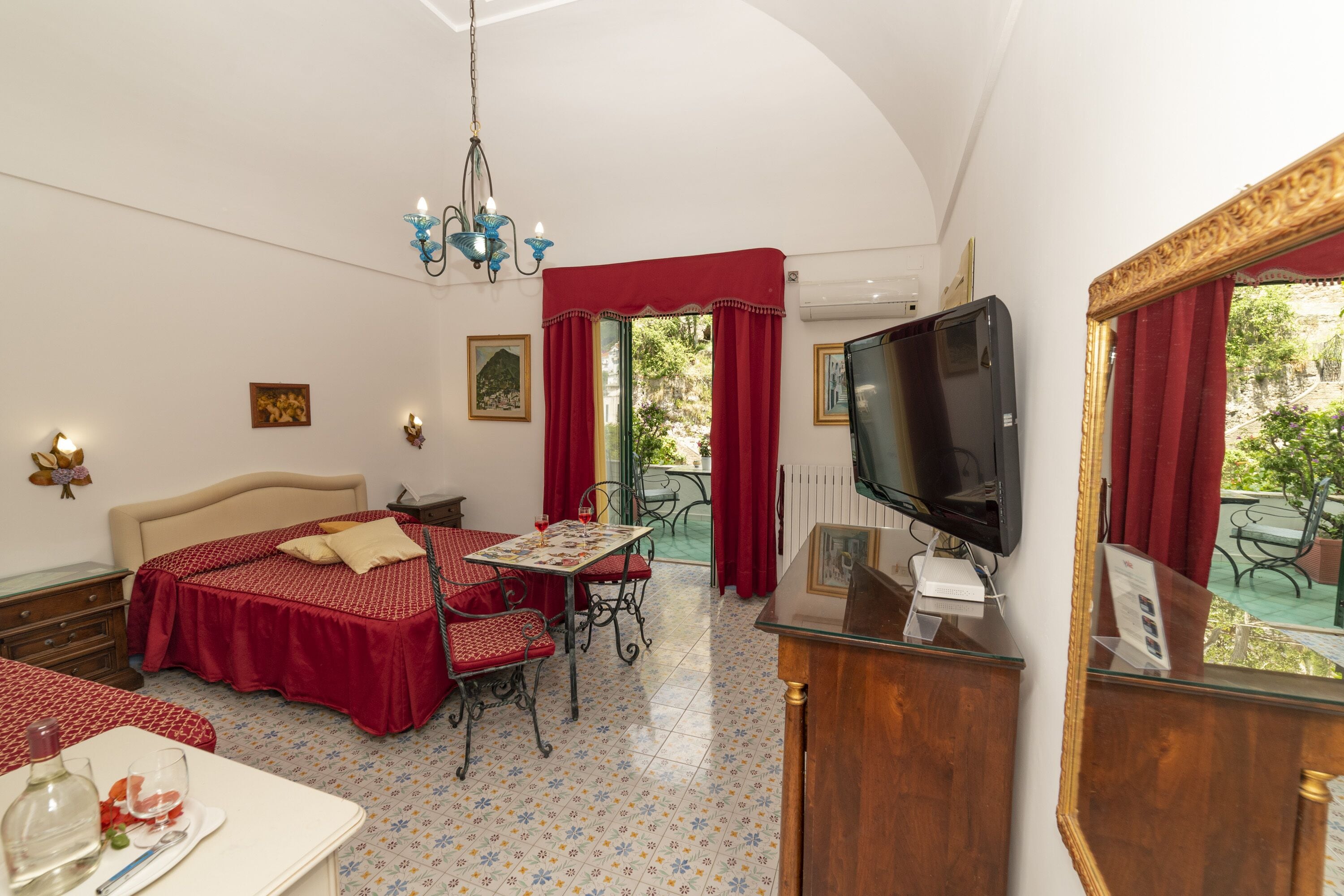 Une chambre d’hôtel avec un lit, une table à manger, des chaises, un téléviseur et un balcon avec des rideaux rouges.