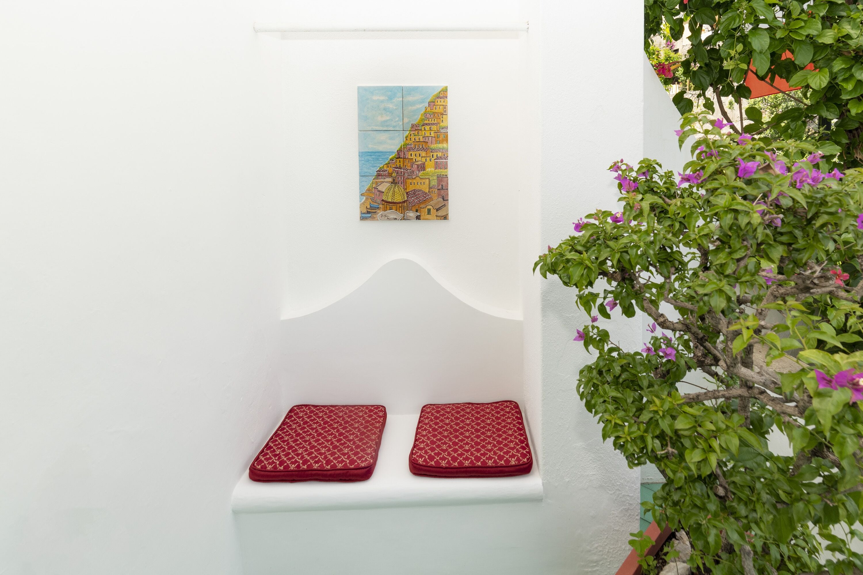 A white wall with a painting of a coastal town, a small bench with red cushions, and a potted plant with green leaves and purple flowers.