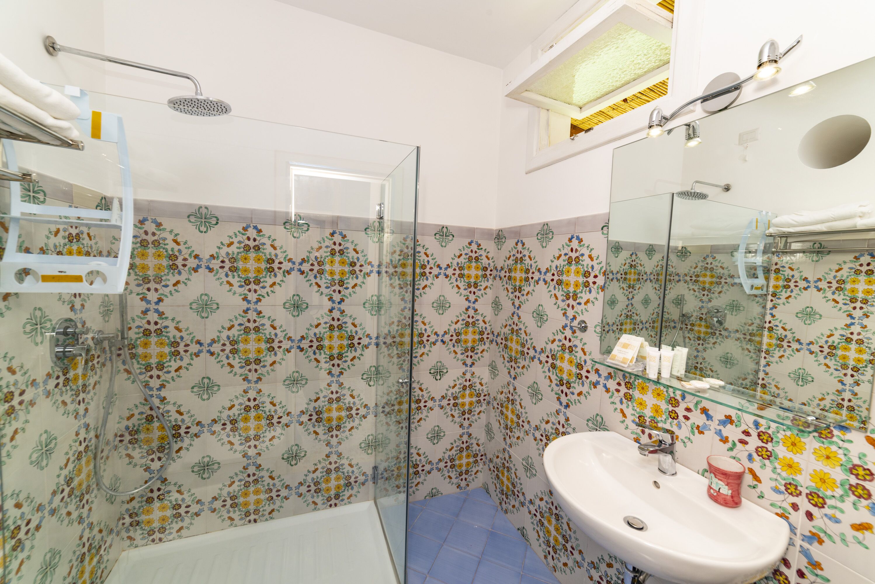 A bathroom with a glass shower enclosure, a white sink, and a mirror with a glass shelf above it.