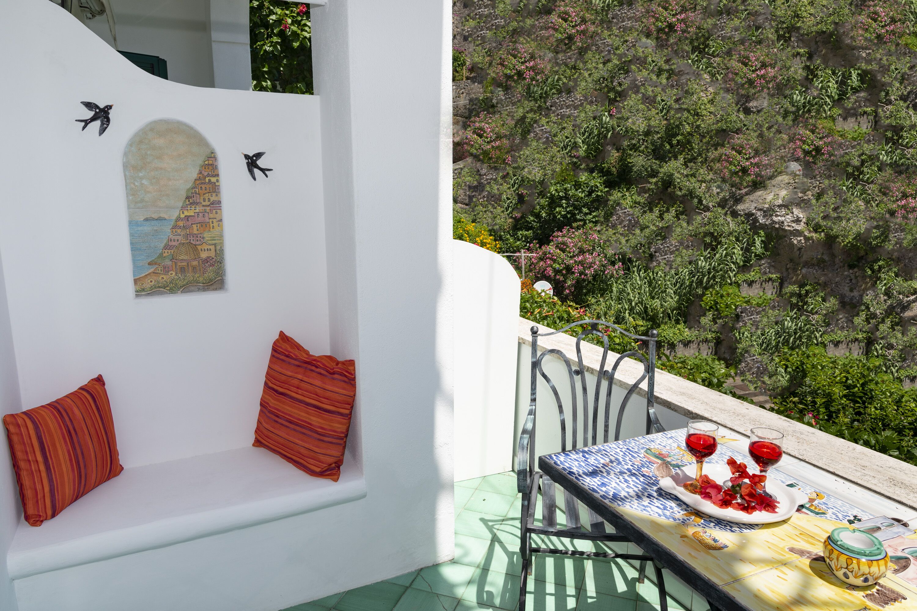Un balcón con pared blanca, un banco con cojines rojos, una mesa con mantel azul y amarillo, dos copas de vino tinto y un plato con comida y flores.