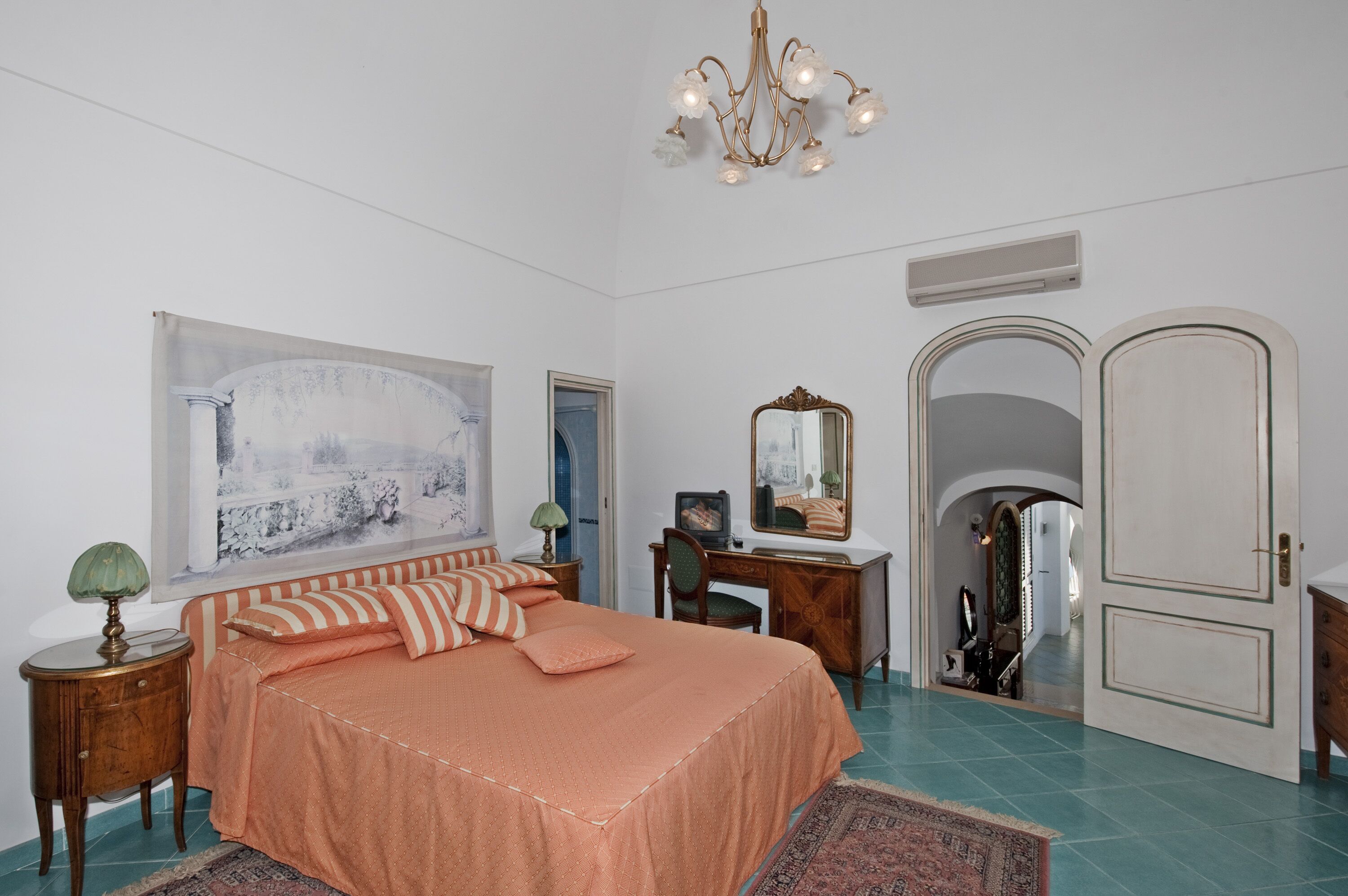A bedroom with a bed, a desk, a chair, a mirror, and a door leading to another room.