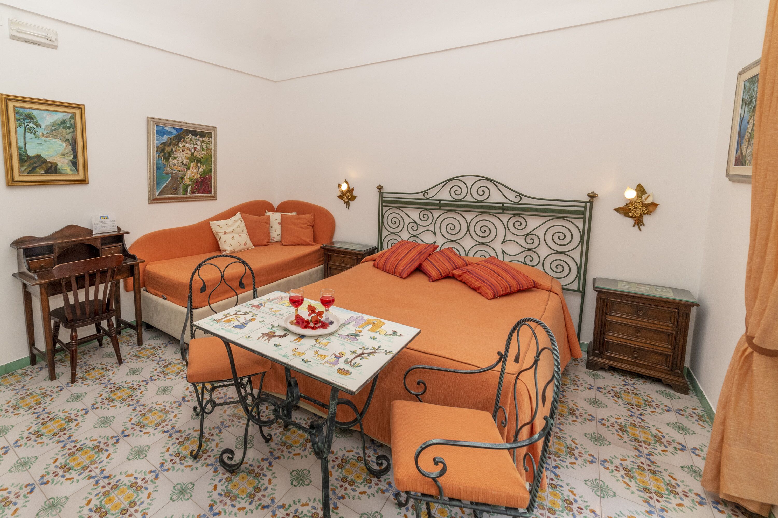 A bedroom with a bed, a small table with drinks, a chair, a nightstand, and two paintings on the wall.