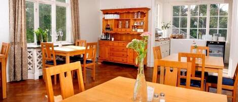 A dining area with wooden tables and chairs, a wooden cabinet with jars, and a window with curtains.