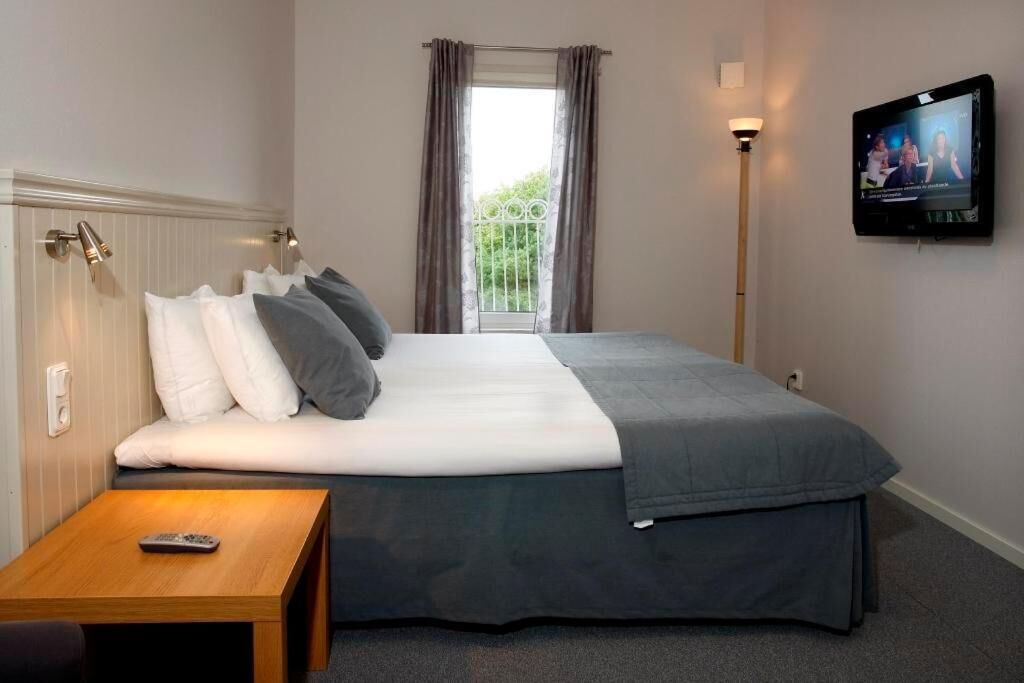 A hotel room with a bed, a TV mounted on the wall, a bedside table, and a window with curtains.