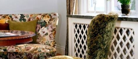 Una habitación con un sofá estampado de flores, un sillón con diseño, suelo de madera, un radiador blanco y una ventana con cortinas.