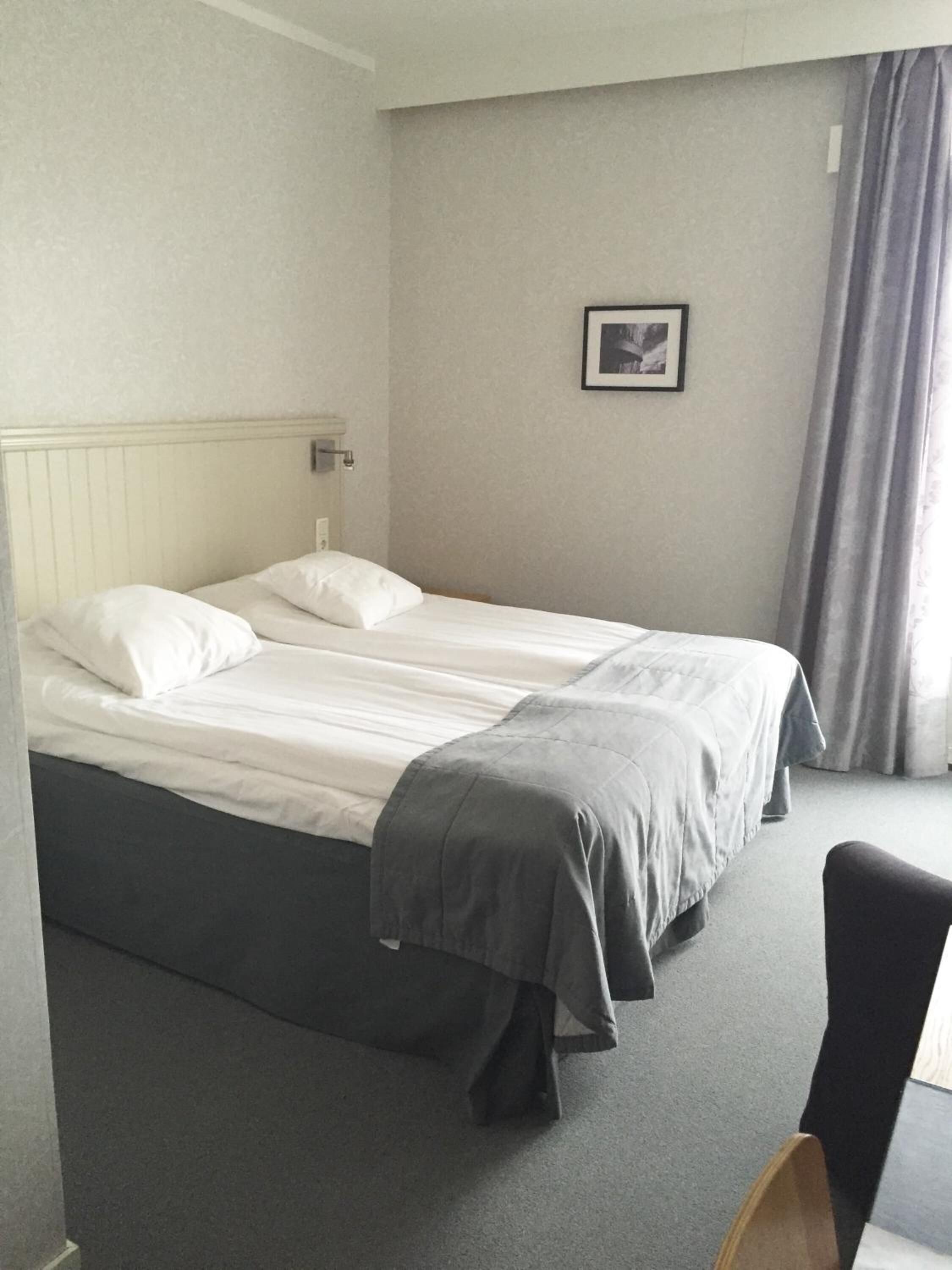 A double bed with white linens and a grey blanket, a bedside table with a lamp, a framed picture on the wall, and a window with curtains.