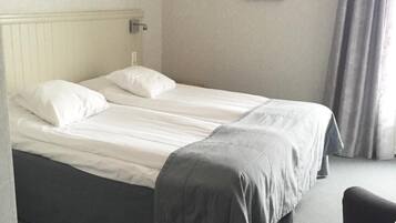 A double bed with white linens and a grey blanket, a bedside table with a lamp, a framed picture on the wall, and a window with curtains.