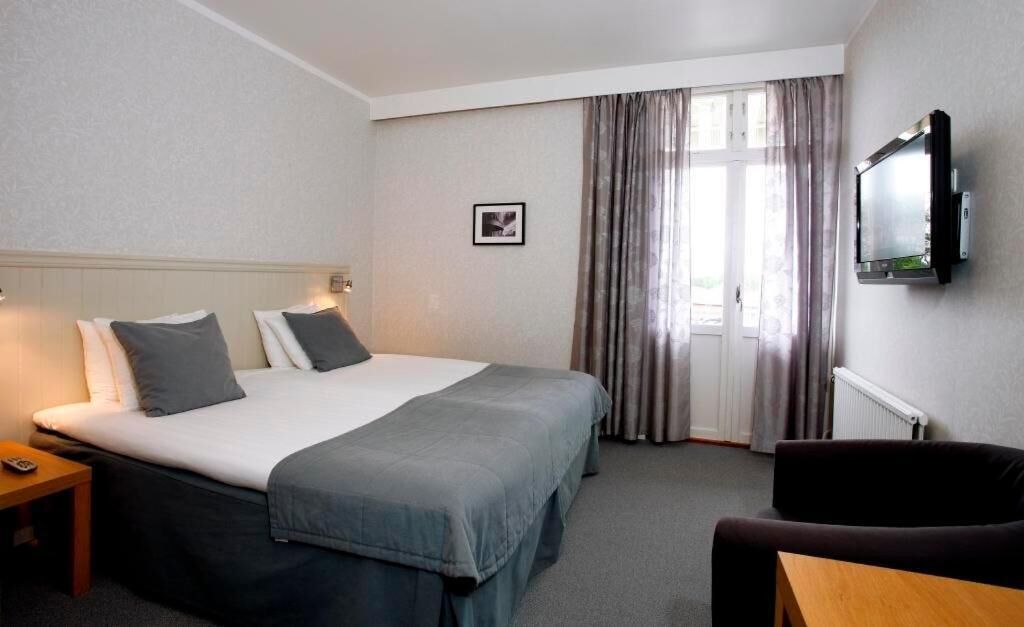 A hotel room with a bed, a TV mounted on the wall, a window with curtains, and a small table.