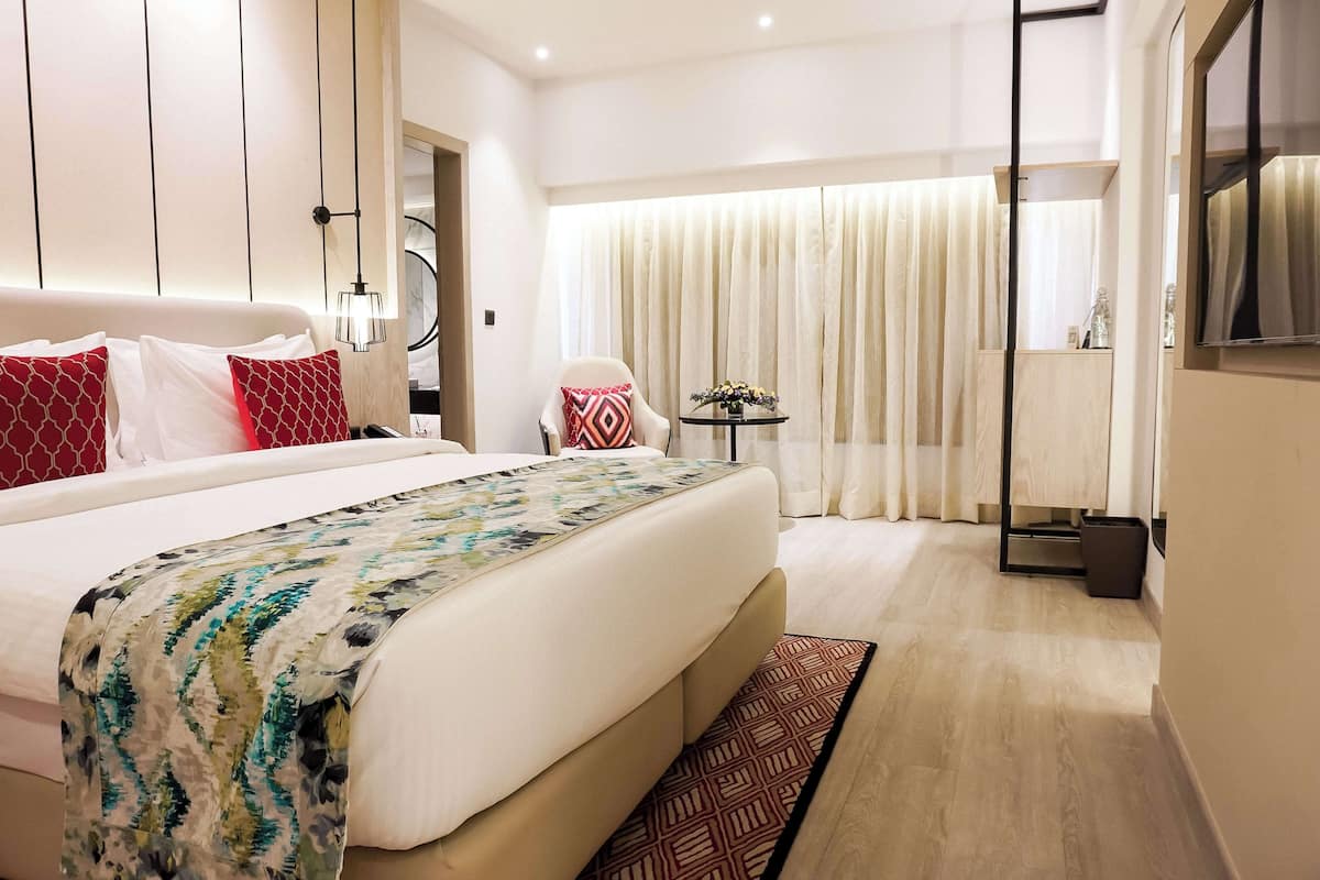 Une chambre d’hôtel avec un grand lit, une table de chevet, une chaise, une petite table avec des fleurs et une grande fenêtre avec des rideaux.