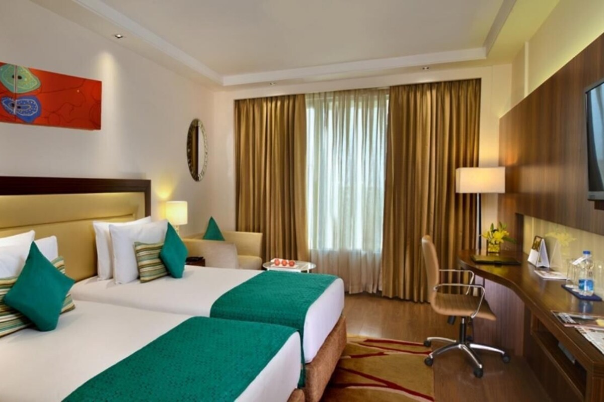 Habitación de hotel con dos camas, un escritorio, una silla, un televisor y un ventanal con cortinas.