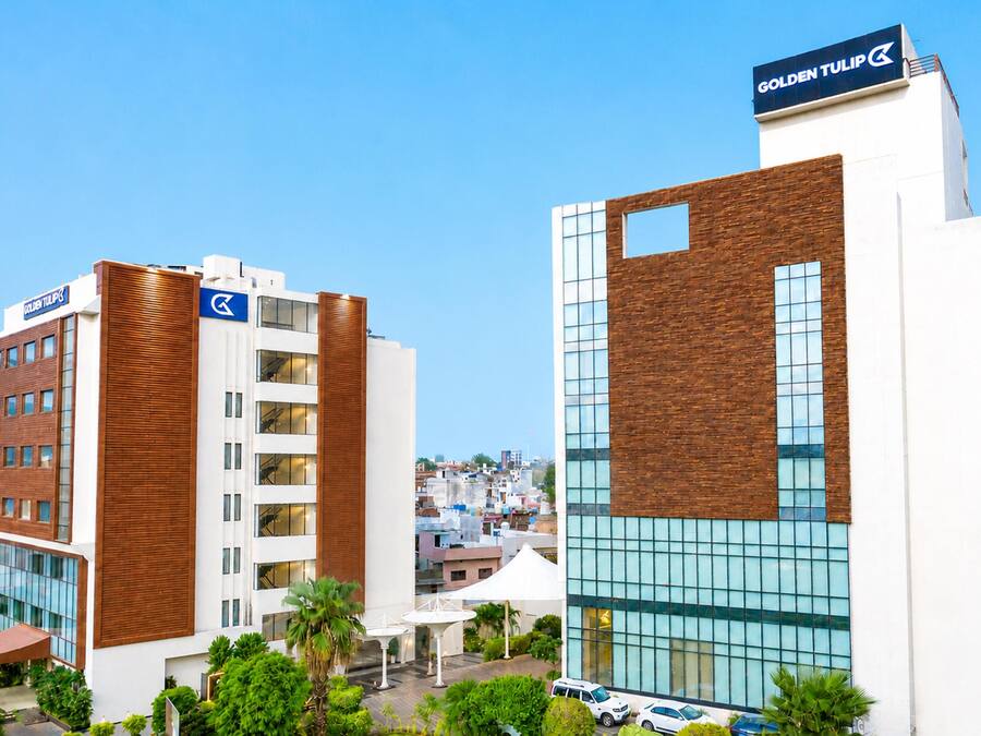 A modern hotel with a prominent 'Golden Tulip' sign, a rooftop terrace with palm trees, and a view of the cityscape.