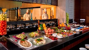 Un buffet proposant une variété de plats et de boissons, des chefs qui s’affairent en arrière-plan et un espace cuisine.