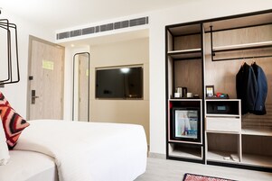 Una camera d'albergo moderna con un letto bianco, un armadio pieno di vestiti, un piccolo frigorifero e una finestra.