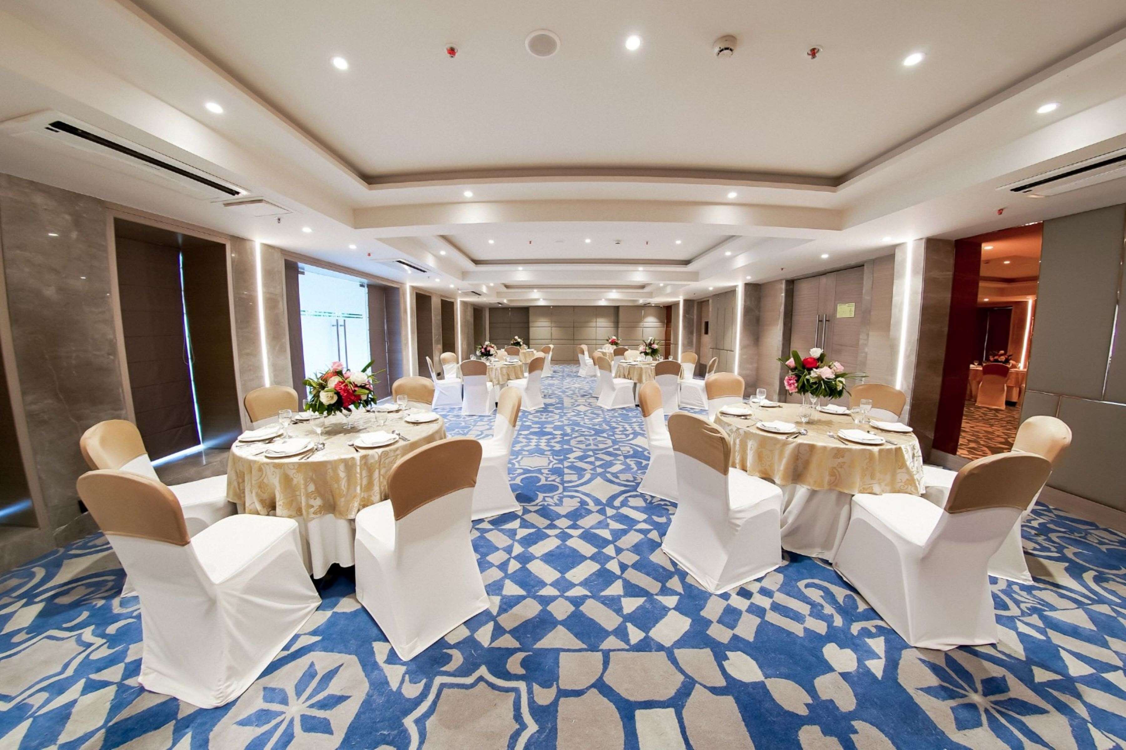 A large, well-lit conference room with round tables and white chairs, a patterned carpet, and a modern ceiling design.