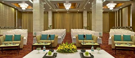 Un salón de conferencias con filas de sillas blancas, dos sofás con cojines color turquesa y una mesa con botellas de agua y una manzana verde.