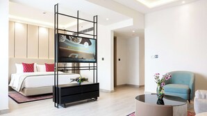 Une chambre d’hôtel moderne avec un lit, un téléviseur fixé sur un support noir, un fauteuil bleu et une table basse ronde ornée d’un vase de fleurs.