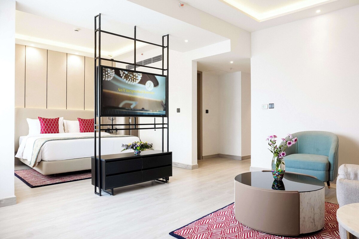 Una habitación de hotel moderna con una cama, un televisor montado sobre un soporte negro, un sillón azul y una mesa de centro redonda con un jarrón de flores.