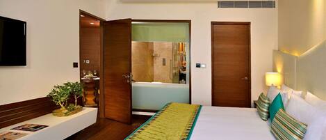 Habitación de hotel con cama, mesita de noche con planta, televisor y baño a la vista por una puerta abierta.