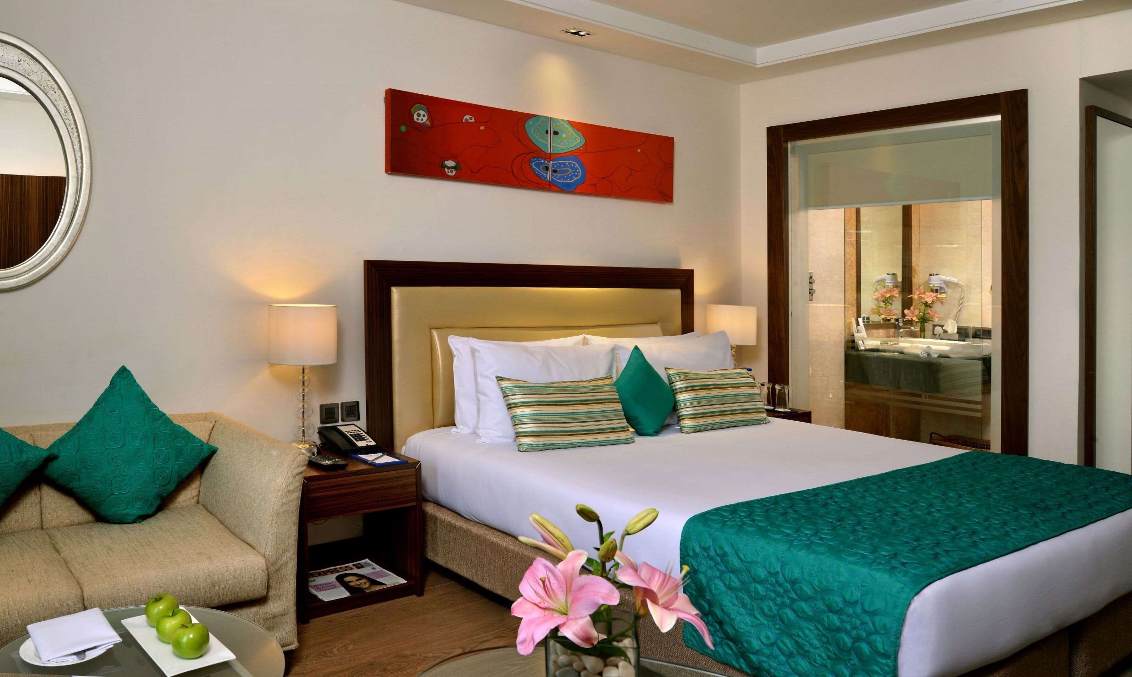 A hotel room with a bed, a sofa, a nightstand with a phone, a mirror, and a painting on the wall.
