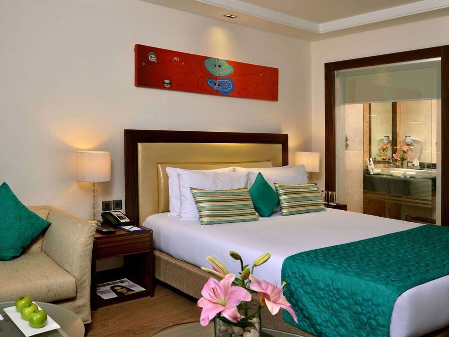 A hotel room with a bed, a sofa, a nightstand with a phone, a mirror, and a painting on the wall.