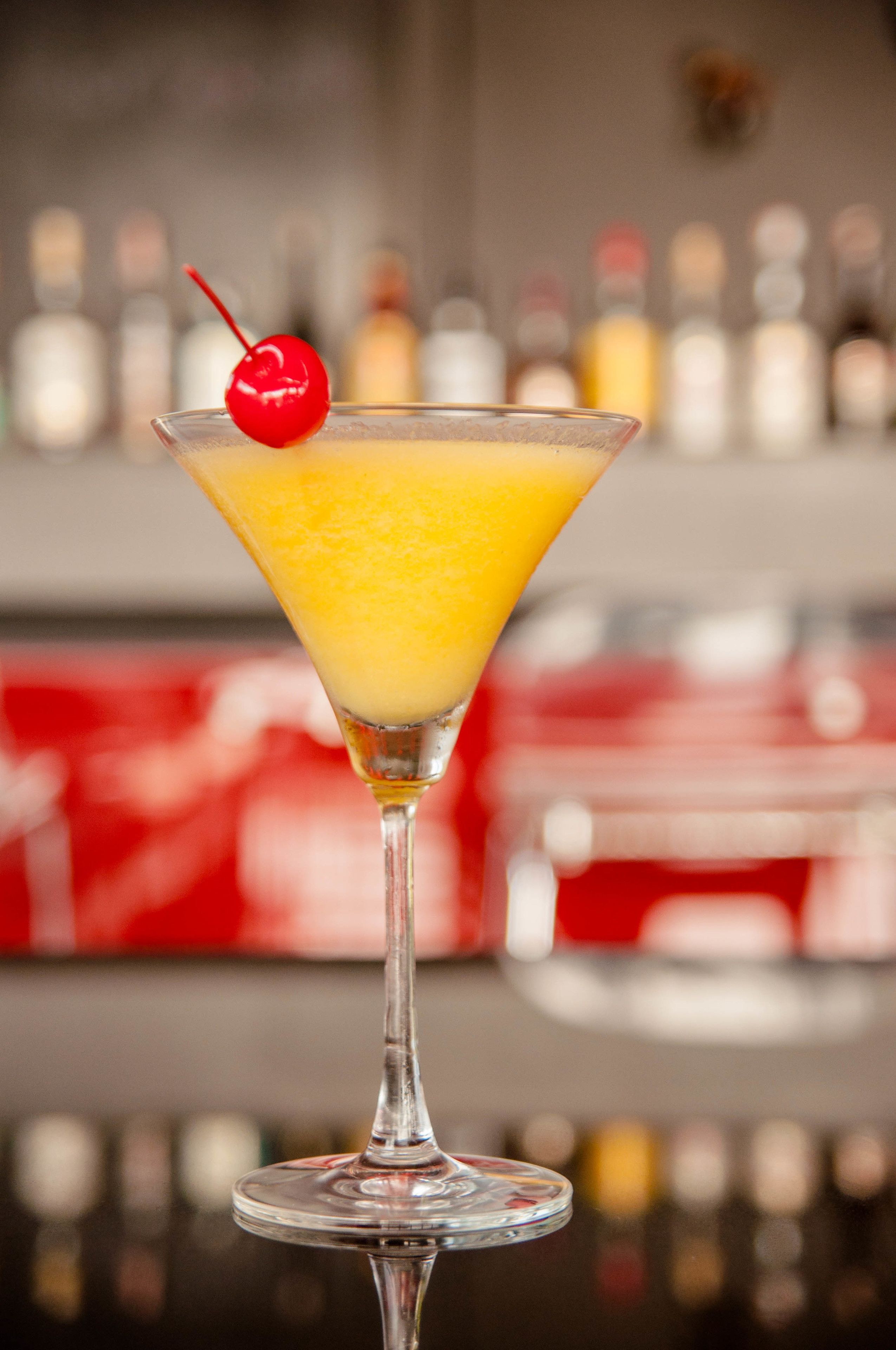 Un got de martini amb un líquid groc i una cirera a sobre, col·locat sobre un taulell de bar amb ampolles al fons.