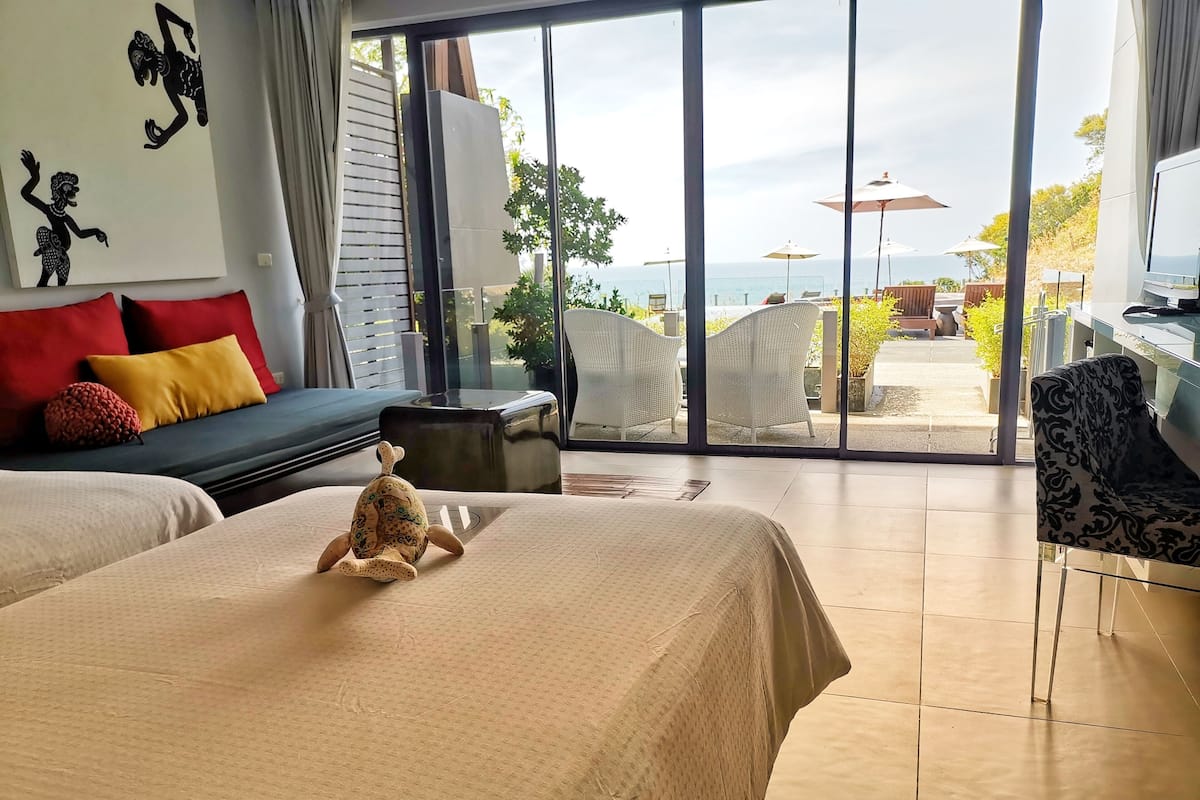 A hotel room with a large bed, a sofa, a small table, and a view of the sea through large windows.