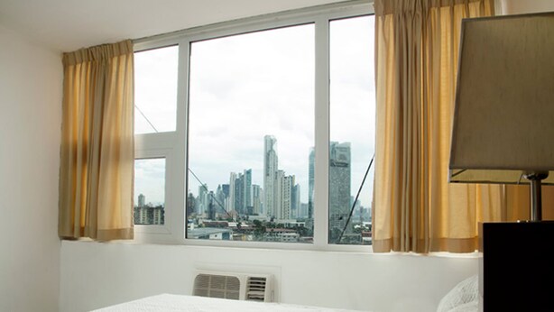 A hotel room with a large bed, a window with curtains, a city view, and a lamp. - Hotel República (Panama City)