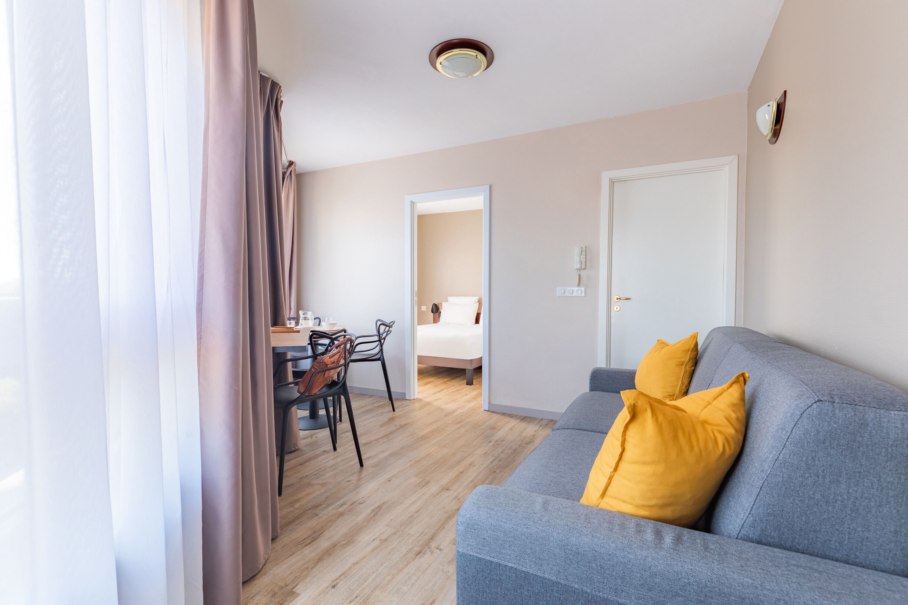 Een moderne woonkamer met een grijze bank, gele kussens, een eethoek met een tafel en stoelen, en een zichtbare deur die leidt naar een slaapkamer.