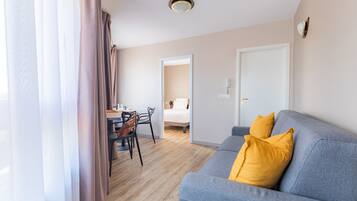 Een moderne woonkamer met een grijze bank, gele kussens, een eethoek met een tafel en stoelen, en een zichtbare deur die leidt naar een slaapkamer.