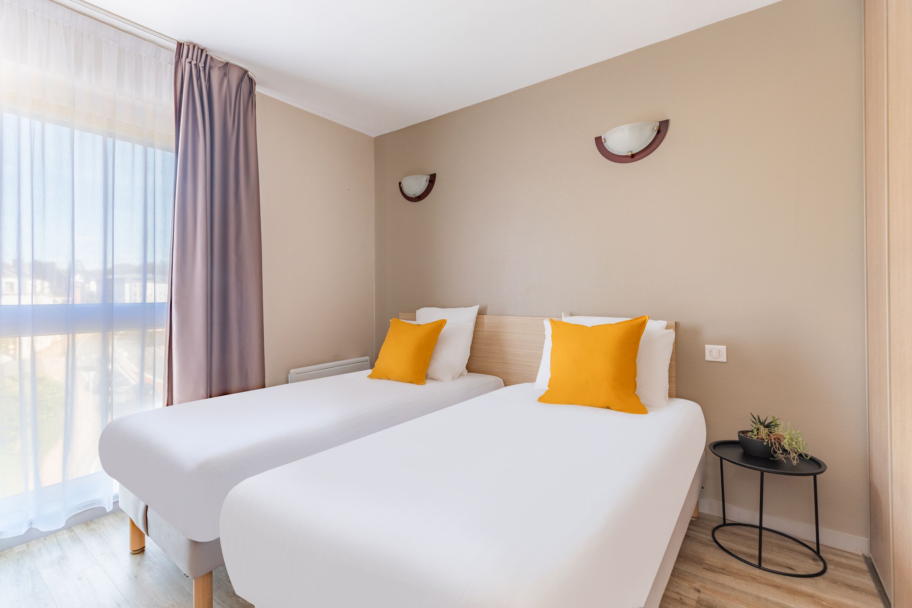 Een hotelkamer met twee bedden, een klein nachtkastje met een plant en uitzicht op de stad door dunne gordijnen.