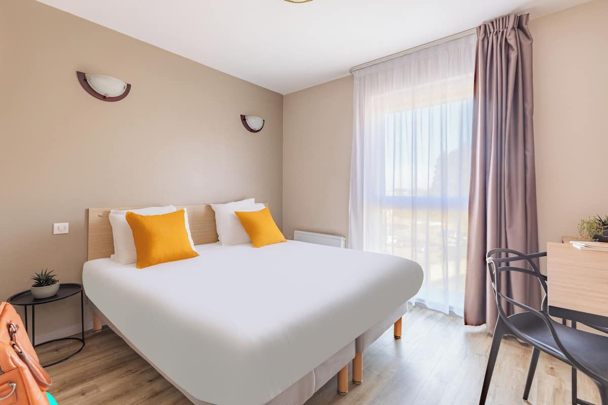 Une chambre d’hôtel avec un grand lit, une petite table, une chaise et une fenêtre avec des rideaux.