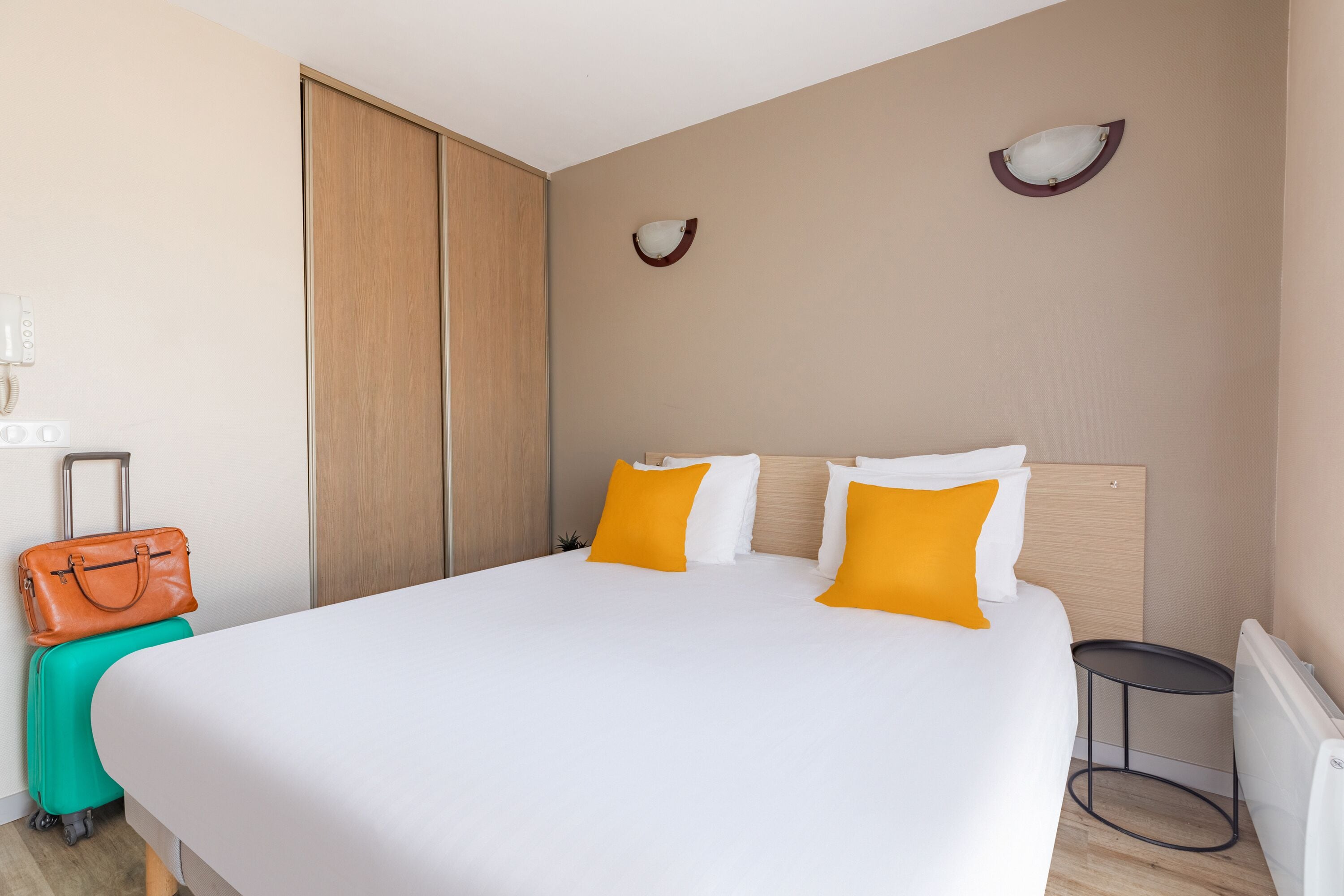 Habitación de hotel con una cama grande, una maleta, una mesita pequeña y una lámpara fijada a la pared.