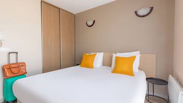 Habitación de hotel con una cama grande, una maleta, una mesita pequeña y una lámpara fijada a la pared.
