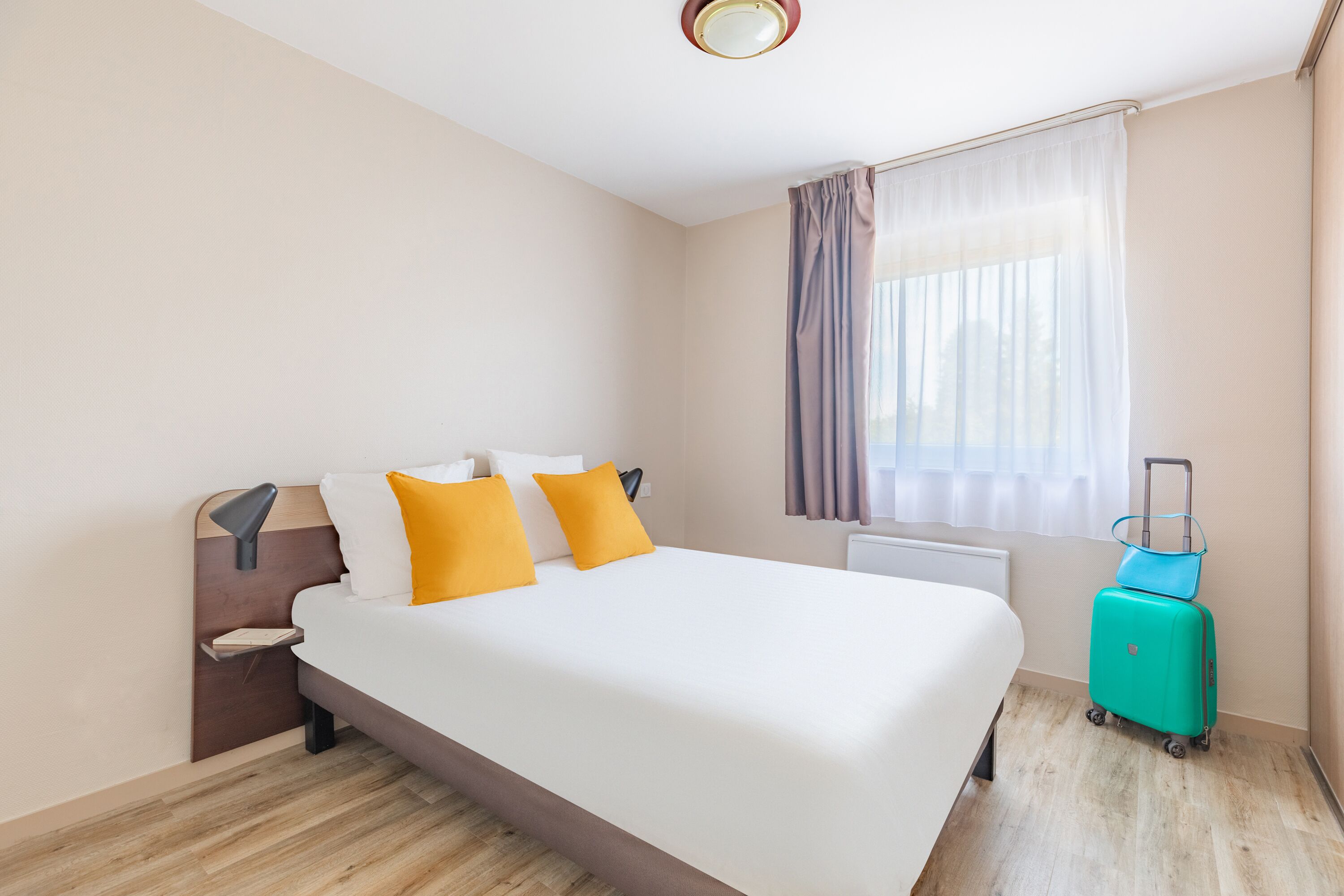 Une chambre d’hôtel avec un lit, une table de chevet, une lampe, une fenêtre avec des rideaux et une valise.