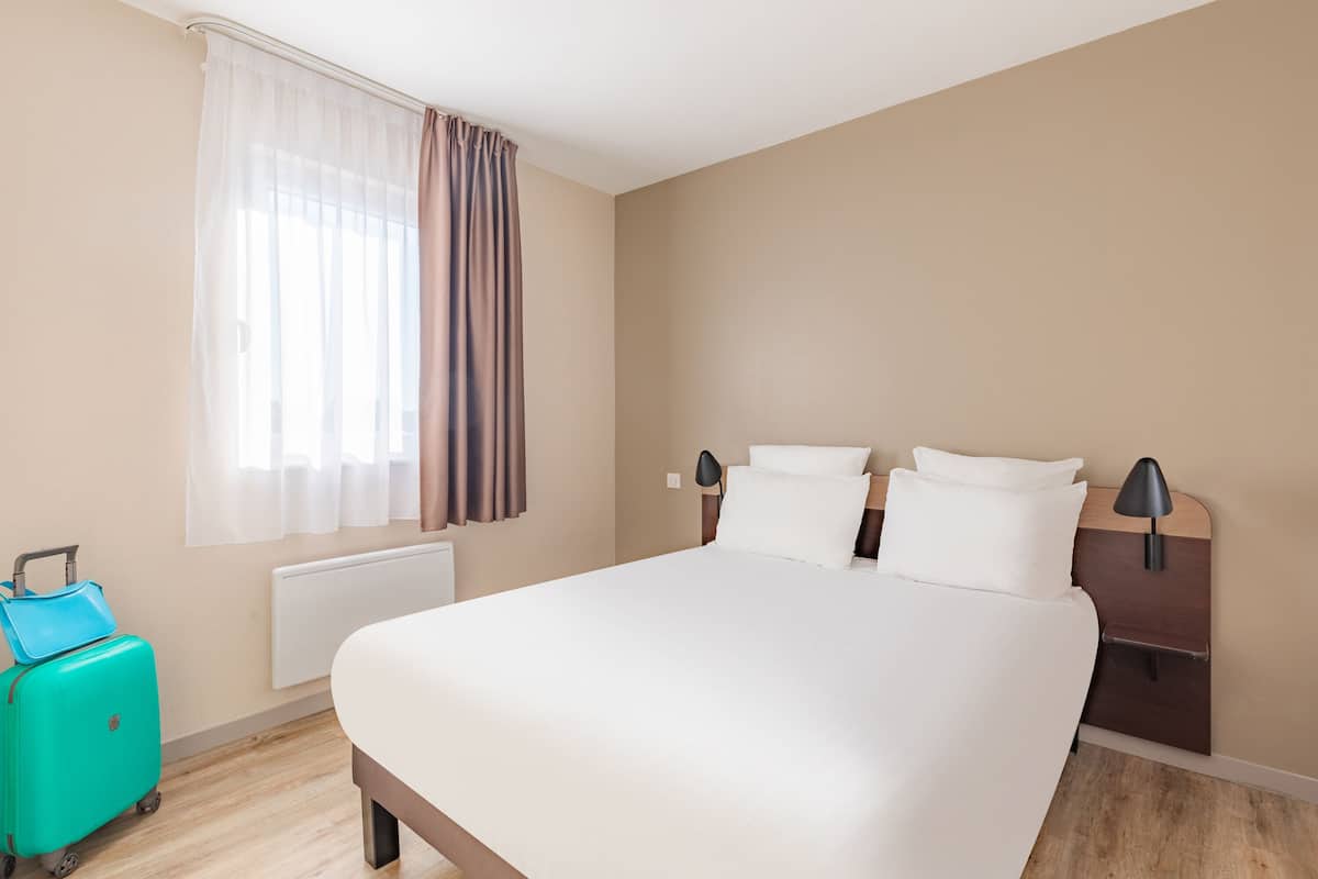 Une chambre d’hôtel avec un grand lit, une table de chevet avec une lampe, une valise et une fenêtre avec des rideaux.
