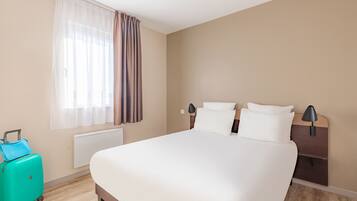 Ein Hotelzimmer mit Bett, Nachttisch, Lampe, einem Fenster mit Vorhängen und einem Koffer.