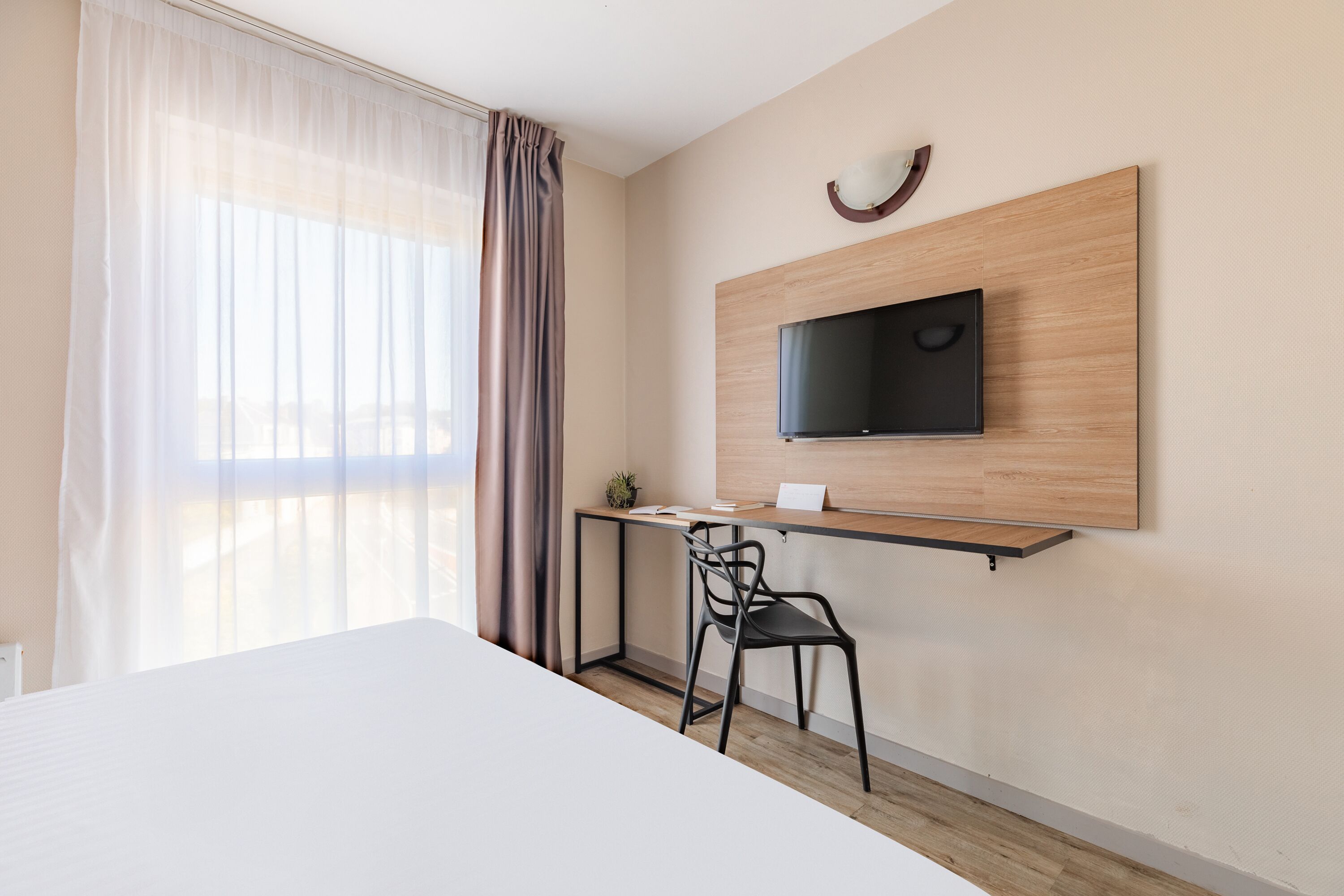 A hotel room with a bed, a desk with a chair, a television mounted on the wall, and a window with curtains.