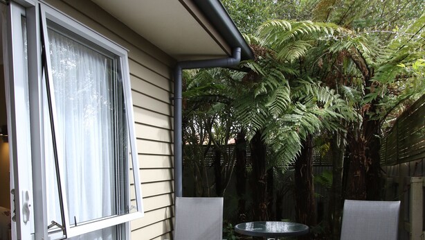 A wooden deck with two chairs and a table, a sliding glass door, and a view of a garden with ferns. - Annabelle Motel (Hokitika)