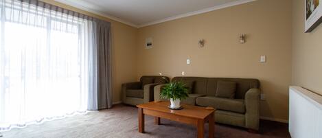 Una sala de estar con un sofá color beige, una mesa de centro de madera con una planta y un ventanal con cortinas.