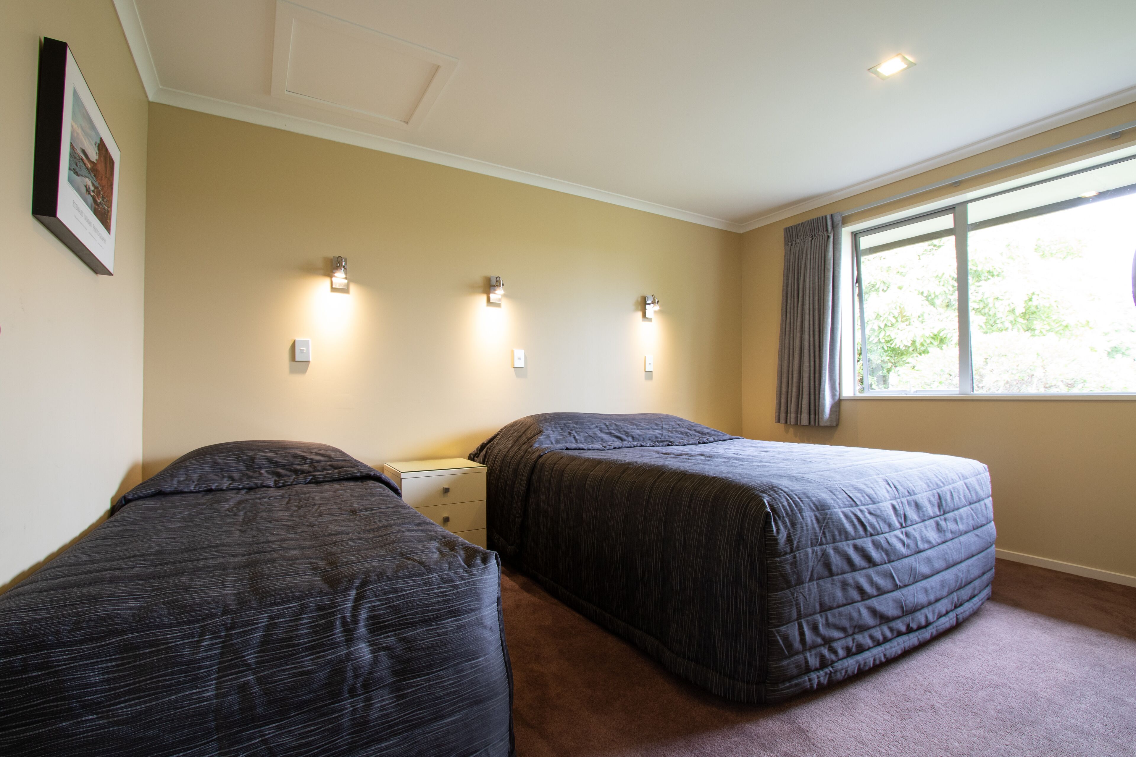 Una habitación de hotel con dos camas individuales, una mesita de noche, una ventana con cortinas y luces fijadas a la pared.