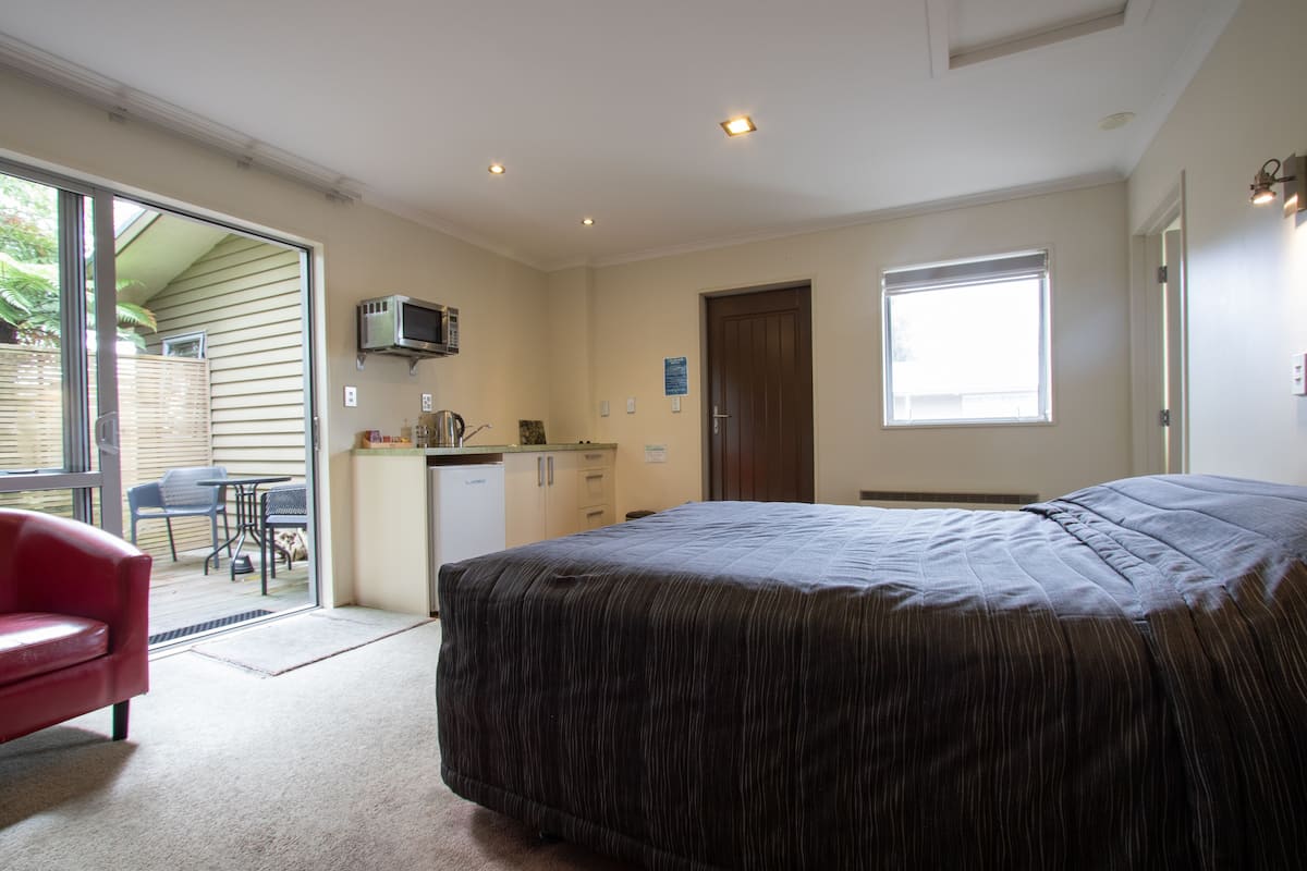 A bedroom with a large bed, a red chair, a kitchenette, and a sliding glass door leading to an outdoor area.