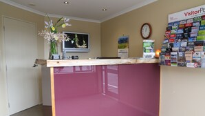 A reception area with a wooden counter, a clock, a wall-mounted TV, and a bulletin board with brochures. - Annabelle Motel (Hokitika)
