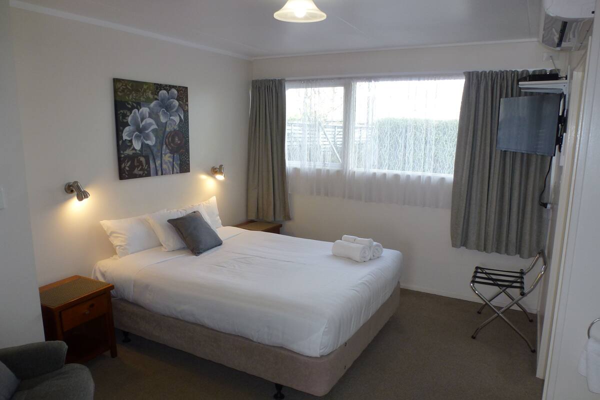 A neatly made bed with white linens, a painting on the wall, a television mounted above the bed, and a window with curtains.
