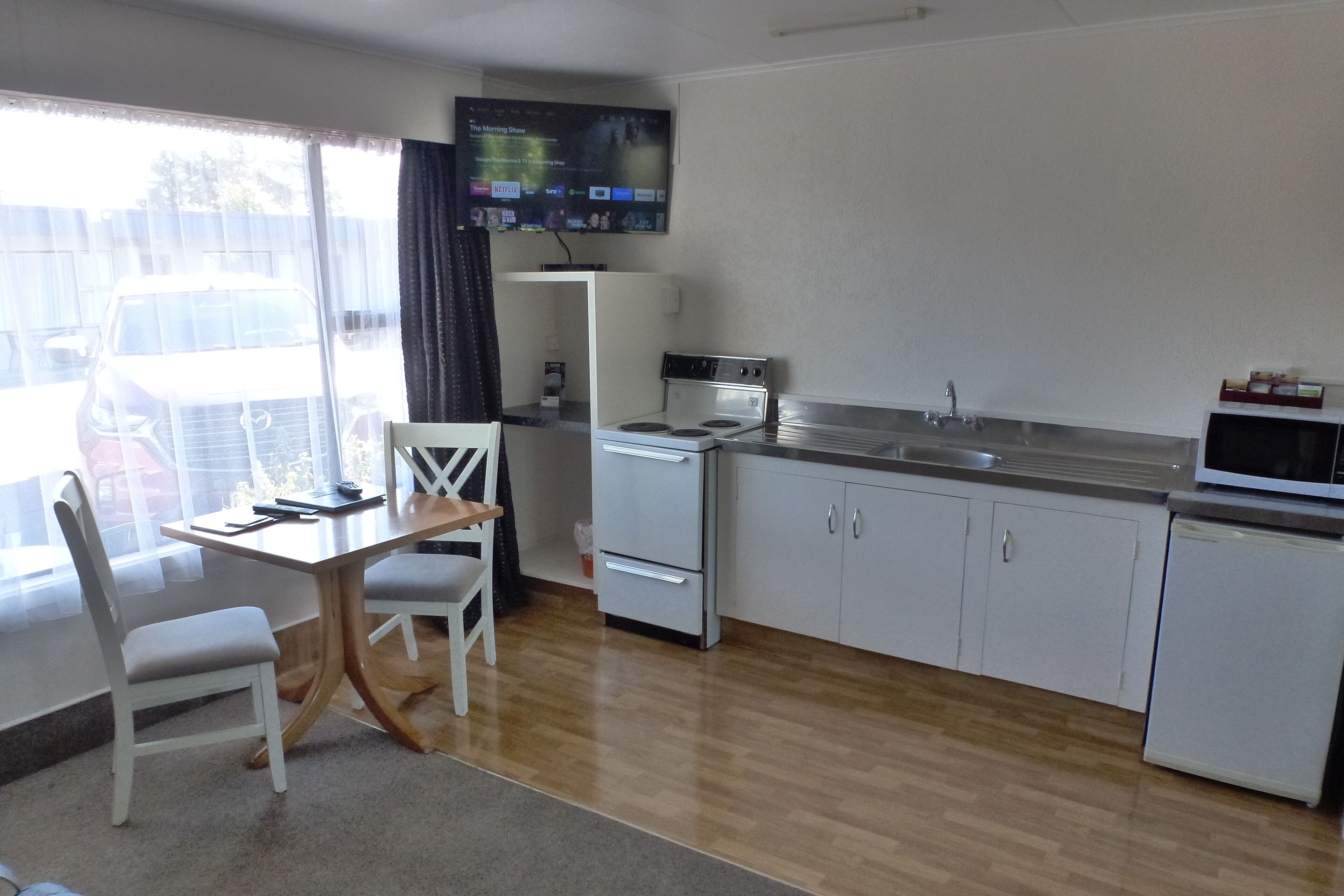 A compact kitchen with a dining area, a TV mounted above the counter, and a view of parked cars outside.