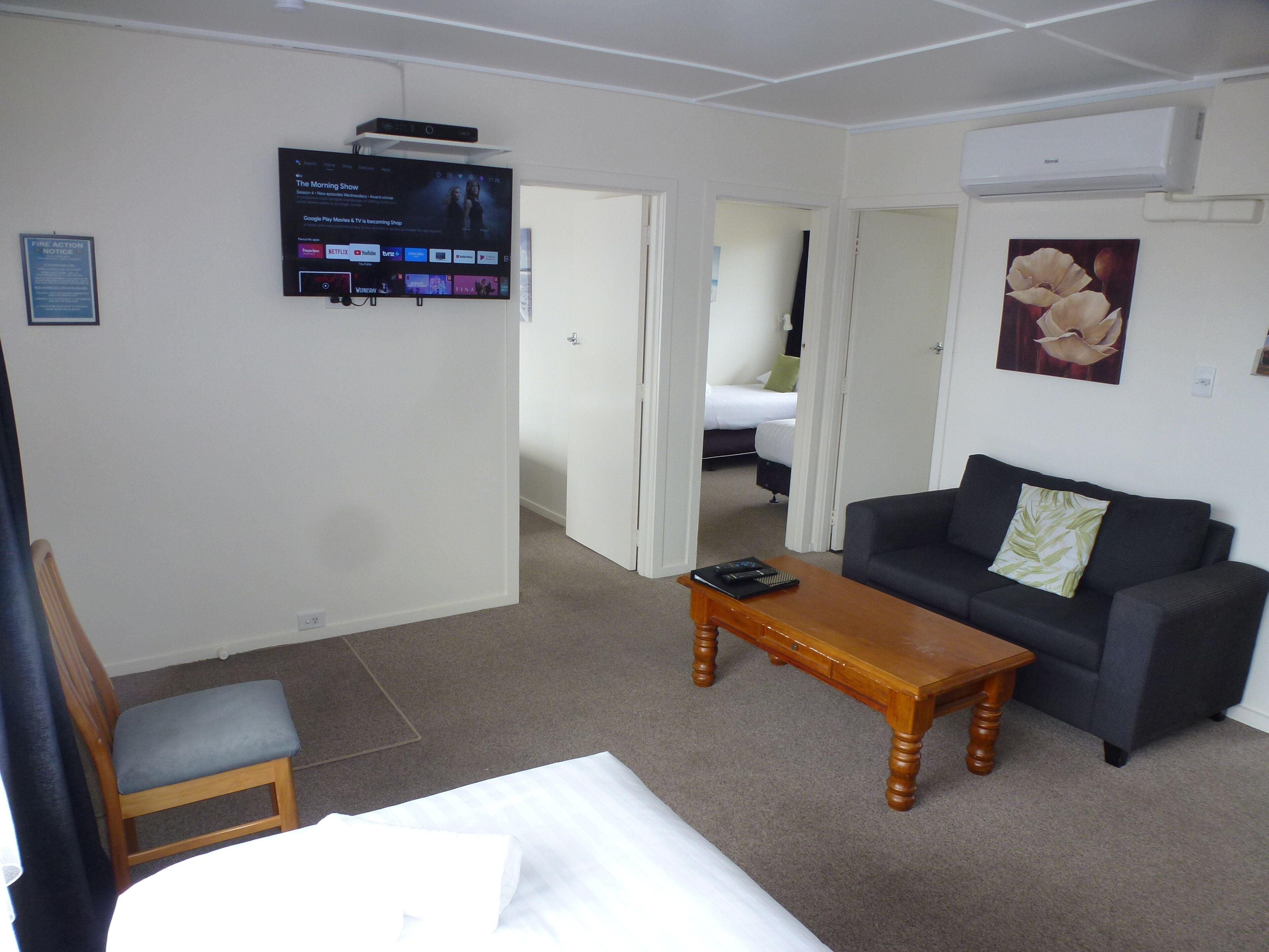 Une chambre d’hôtel avec un lit, une table basse en bois, un canapé gris, un téléviseur fixé au mur et un tableau représentant une fleur.