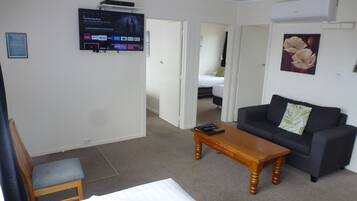 Une chambre d’hôtel avec un lit, une table basse en bois, un canapé gris, un téléviseur fixé au mur et un tableau représentant une fleur.
