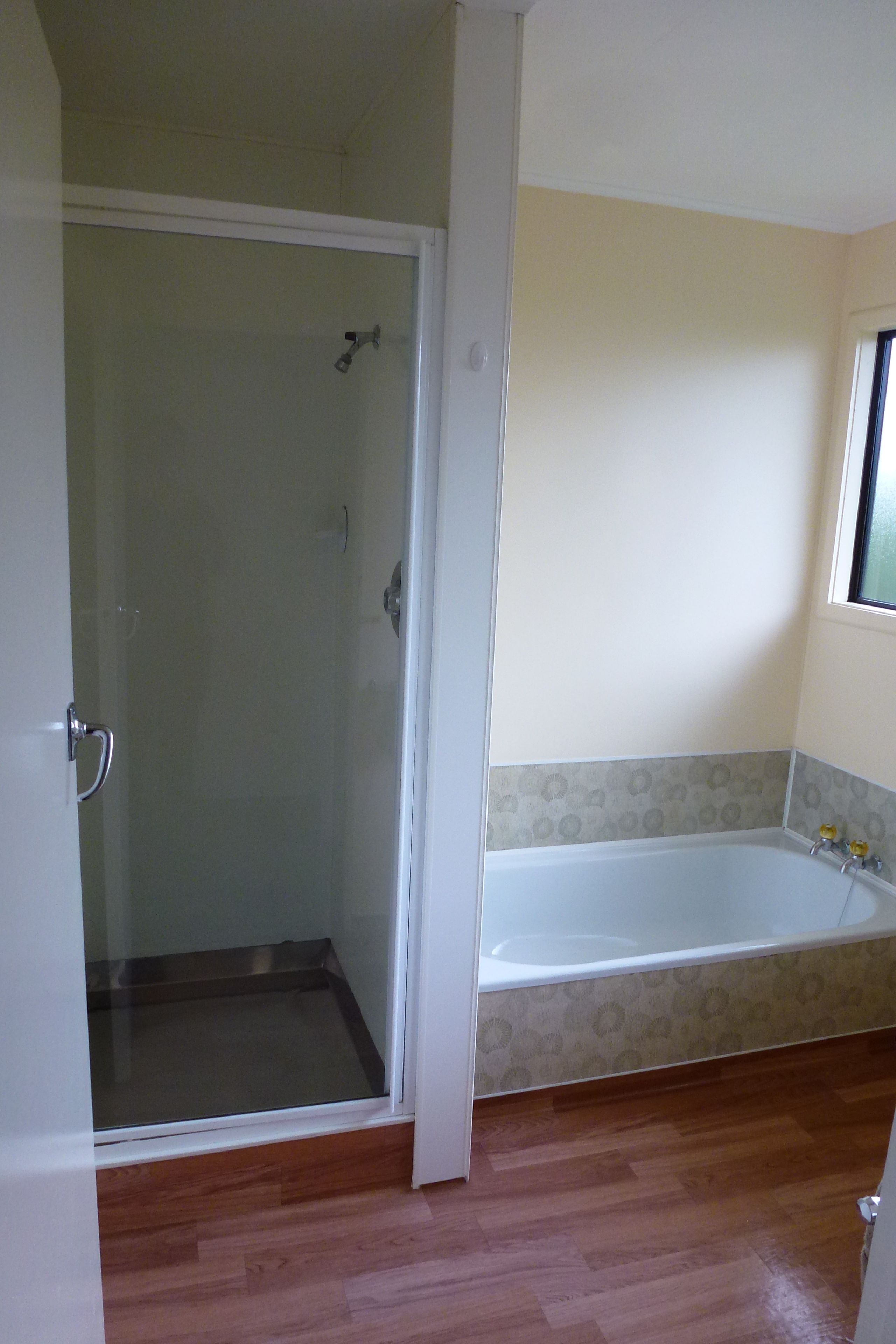 A bathroom with a glass-enclosed shower, a white bathtub with a patterned tile border, and wooden flooring.