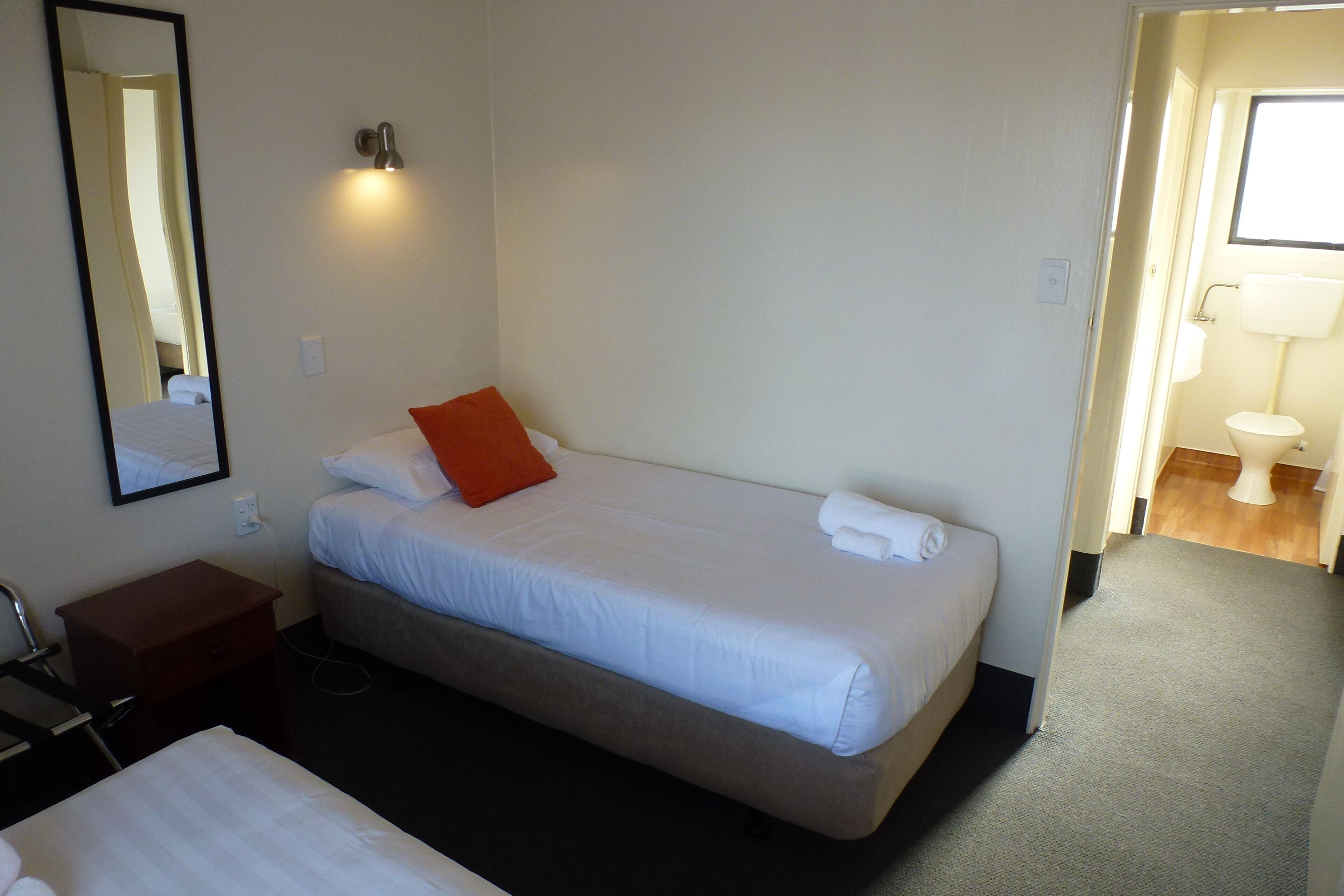 Habitación de hotel con una cama individual, una mesita de noche, un espejo y un baño visible a través de una puerta abierta.